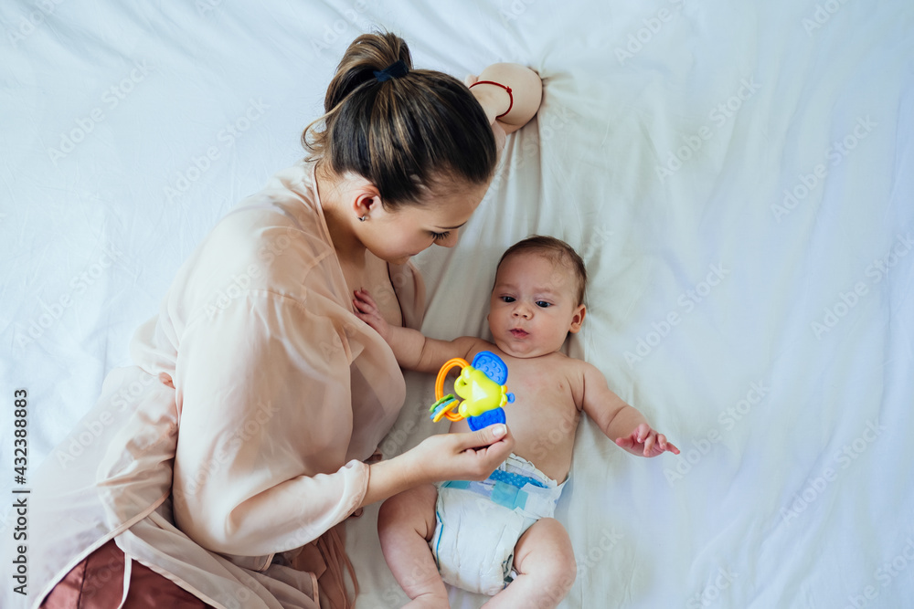 Young Mother Holding Rattle Toy and Lying Down in Bed with her Newborn Baby Single mother and