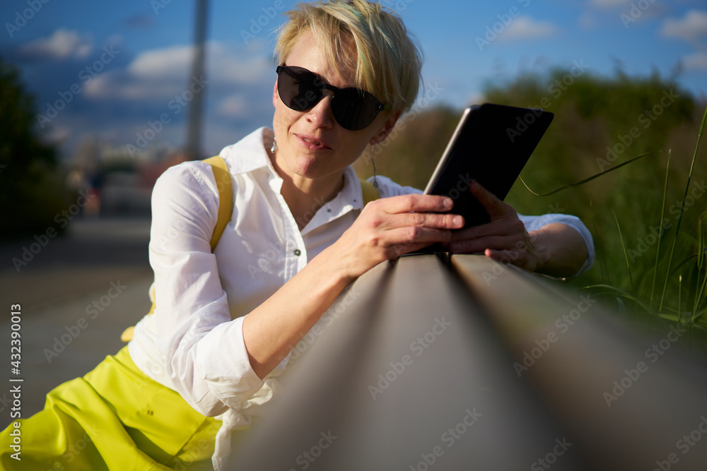 Contemplative woman with short blonde haircut thoughtful looking away resting on bench during sunny day, pondering blogger with modern digital tablet for chatting connected to 4g wireless thinking