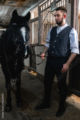 A young man in the guise of a brutal english gangster stands in a stable next to a horse