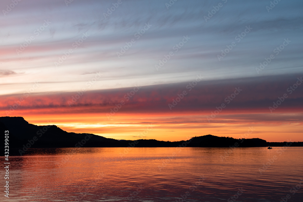 Dramatic seascape, stunning view of sunset at sea with lonely fishing boat in the calm sea reflection under a colorful and vibrant sky with mountain and hill silhouettes at the background. Twilight