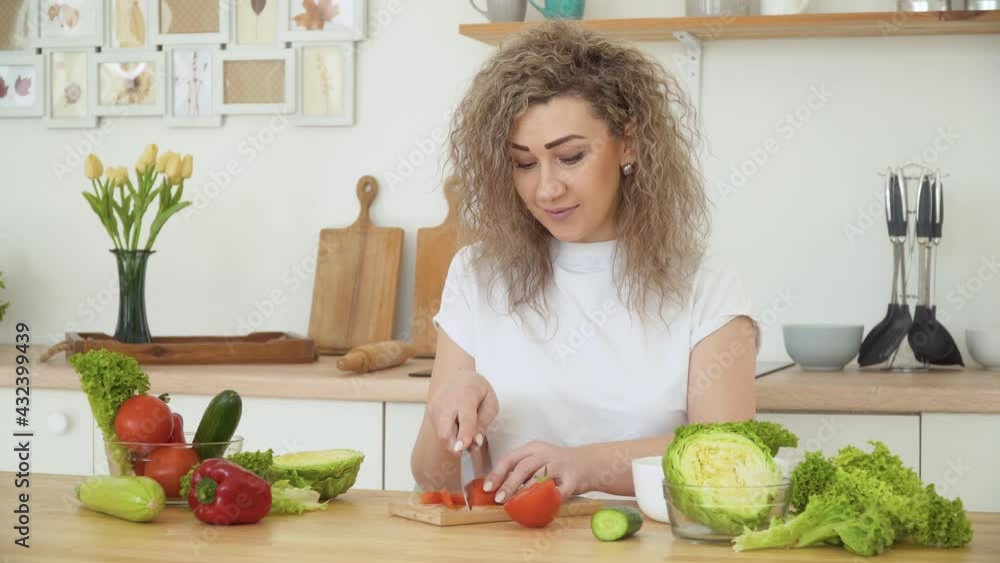 Young blonde woman with curly hair slices a red tomato sitting at a table in the kitchen in a Scandinavian Hygge design. Healthy food and diet concept Young blonde woman with curly hair slices a red tomato sitting at a table in the kitchen in a Scandinavian Hygge design. Healthy food and diet concept