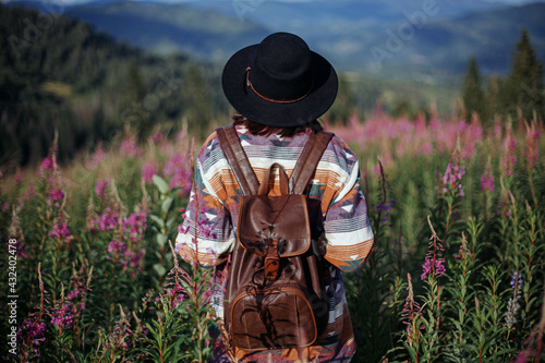 Wanderlust. Woman traveler with backpack and hat standing in wildflowers and looking on mountains
