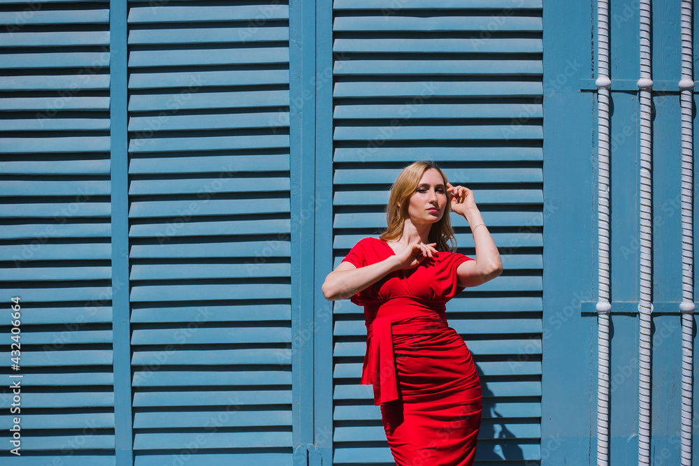 Fototapeta premium A young woman stands near a wall with blue shutters. Bright sunny day, hard light.