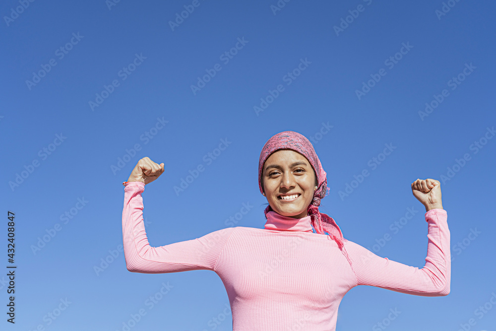 Young woman with cancer showing strength with her smile and arms raised, light blue background and copy space. Hope, Struggle, Empowerment Concept. Young woman with cancer showing strength with her smile and arms raised, light blue background and copy space. Hope, Struggle, Empowerment Concept.