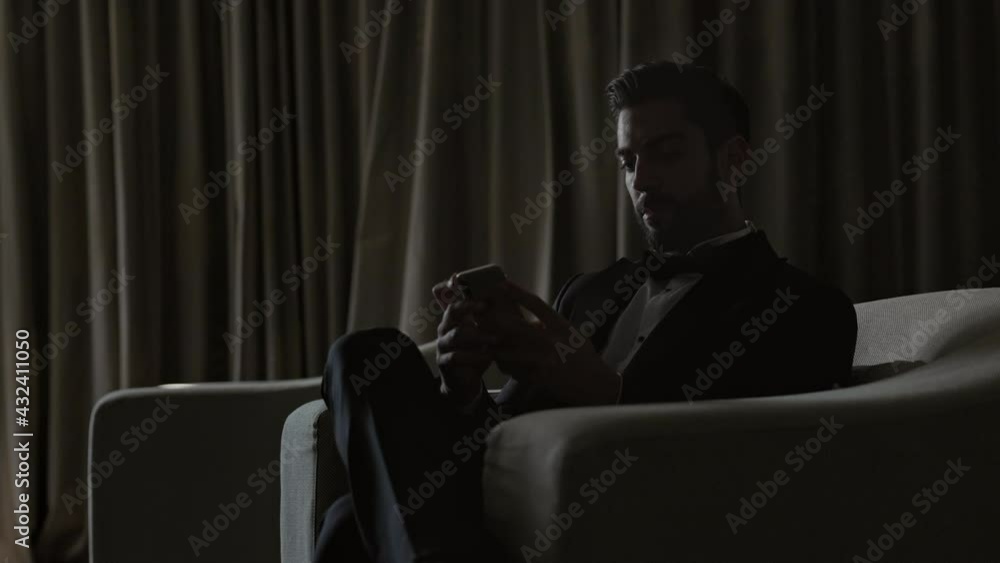 Stylish Young Man Sitting On Sofa With Cellphone In Hands. Man In Suit And White Shirt. In The Room Is Dark. Stylish Young Man Sitting On Sofa With Cellphone In Hands. Man In Suit And White Shirt. In The Room Is Dark.