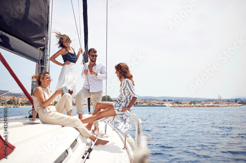A group of young handsome models is relaxing with a coffee on the deck of the yacht and chatting while riding through the dock on the seaside. Summer, sea, vacation, friendship