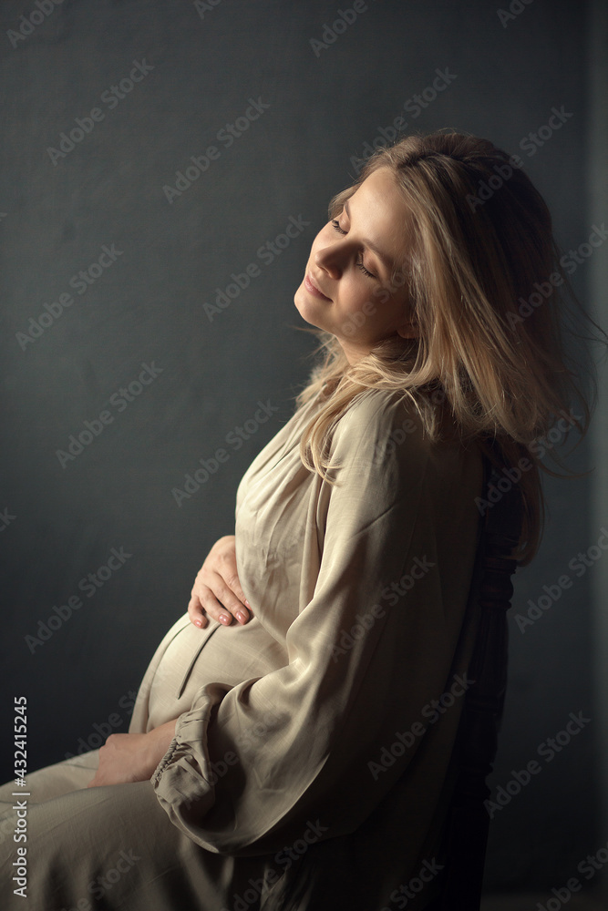 Portrait of a beautiful pregnant young woman sitting on a chair in the dark room. Image with selective focus and toning. Portrait of a beautiful pregnant young woman sitting on a chair in the dark room. Image with selective focus and toning.