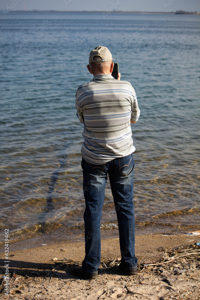 Elderly man stands with his back and photographs the sea 