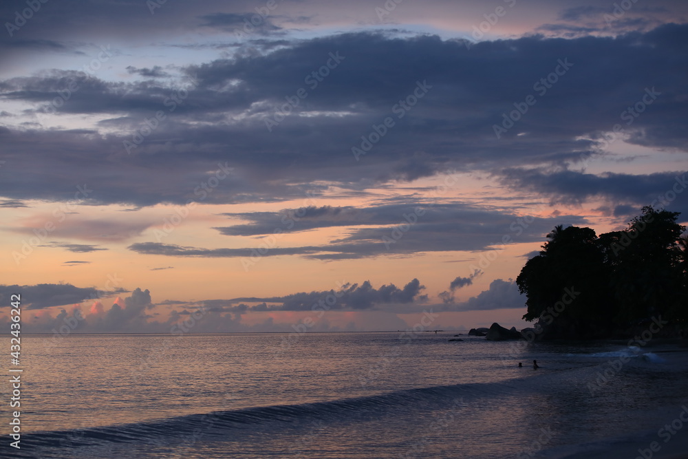 Pink sea sunset with calm water and gray clouds on the horizon. Image of a tropical evening with people swimming in the distance
