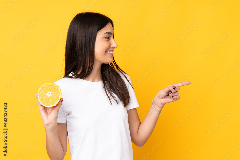 Young caucasian woman holding an orange isolated on yellow background pointing to the side to present a product Young caucasian woman holding an orange isolated on yellow background pointing to the side to present a product