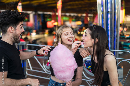 Dos chicas y un chico amigos sonriendo y compartiendo un algodón de azúcar rosa en zona de atracciones de una feria