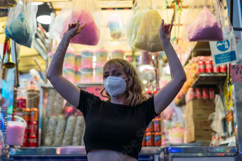 Chica rubia joven con mascarilla posando delante de puesto de algodón de azúcar de una feria