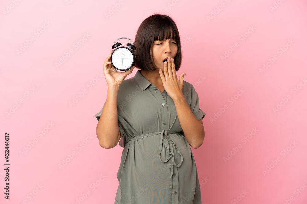 Fototapeta premium Woman over isolated background pregnant and holding clock while yawning