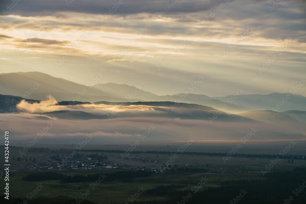 Scenic mountain landscape with orange lilac low clouds above village among mountains silhouettes under dawn cloudy sky. Atmospheric alpine scenery of countryside in low clouds in sundown golden color.