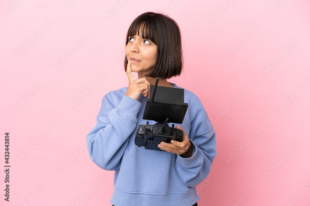Young mixed race woman holding a drone remote control isolated on pink background having doubts while looking up Young mixed race woman holding a drone remote control isolated on pink background having doubts while looking up