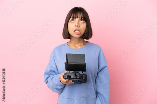 Young mixed race woman holding a drone remote control isolated on pink background looking up and with surprised expression