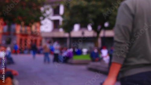 Blurred, unrecognisable people walking around in a city with other people wearing face masks sitting alongside perches, typical city life background of South America