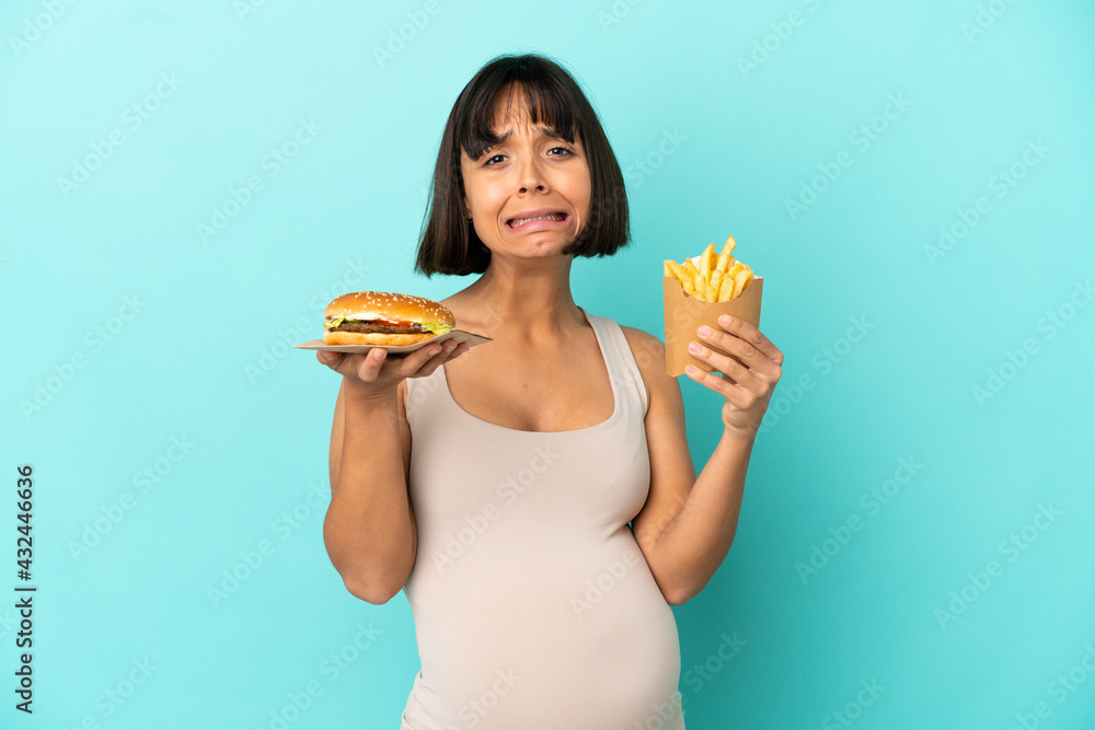 Young pregnant woman holding burger and fried chips over isolated blue background Young pregnant woman holding burger and fried chips over isolated blue background