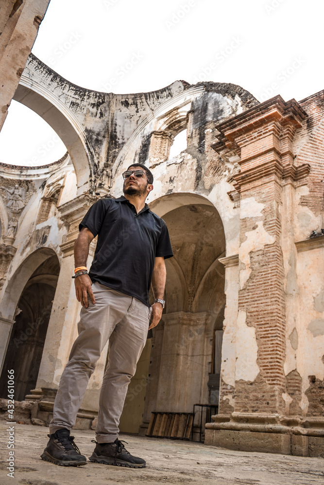 adult man in black shirt and glasses observing the ruins of a cathedral with a classic colonial architecture style with rubble and stones and a dark gray sky
