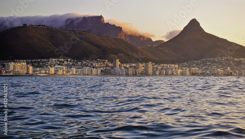View of Cape Town from the Atlantic Ocean. The clouds on Table Mountain are illuminated by the evening sun. Lion's head rock on the background of the sky. There are many city buildings on the shore.