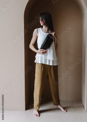 Black Leather Waist Bag For Women Small Leather Bum Bag. Young caucasian woman stands barefoot indoors. The woman is wearing brown pants, a white blouse and a black banana bag.