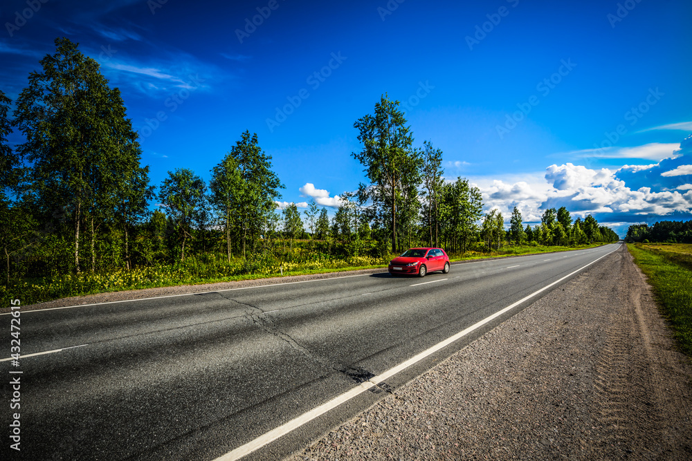 On the road passing through the field and the forest rides a red car. View from the side of the road On the road passing through the field and the forest rides a red car. View from the side of the road