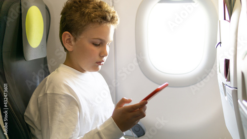 A cute Caucasian teenager sits on a plane near the window and looks at his smartphone.