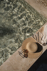 Flat lay of sunglasses and straw hat on marble swimming pool side with clear blue water with waves sunlight shadow reflections. Minimal fashion aesthetic summer vacation top view creative background