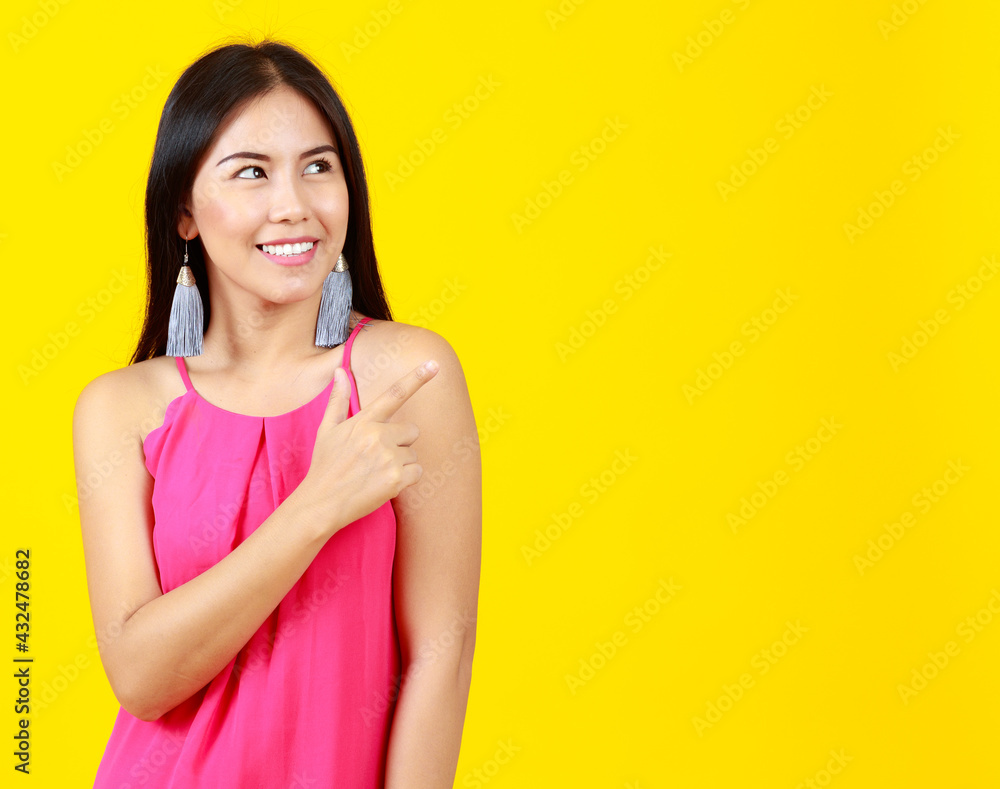 Asian beautiful cheerful adult woman with long black hair wearing pink dress, fashionable earring, wrist watch, happily smiling, pointing to present blank copy space on isolated yellow background. Asian beautiful cheerful adult woman with long black hair wearing pink dress, fashionable earring, wrist watch, happily smiling, pointing to present blank copy space on isolated yellow background.