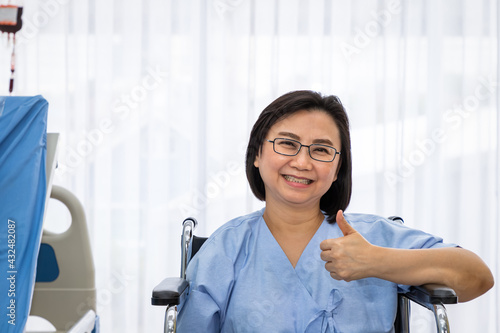 Senior Asian woman patient sitting on wheelchair in wardroom at hospital, thumb-up, treatment and recovering, healthcare concept. photo