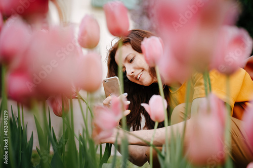 Young smiling girl takes pictures using smartphone of blooming flowers pink tulips in the garden. Allergy-free travel and fragrance concept.