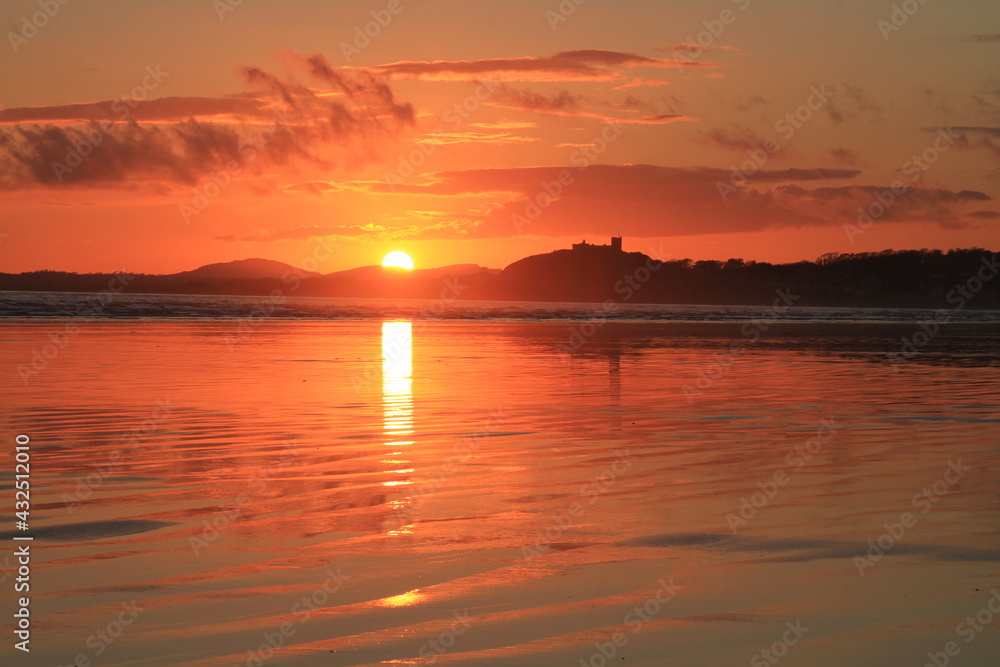 Setting sun drops to the horizon, silouetting Criccieth Castle, and casts reflections across the rippled sand at Black Rock Beach (landscape).