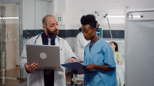 African nurse and surgeon doctor in medical uniform analyzing illness symptom working in hospital ward. In background woman physician checking sick patient while recovering after surgery