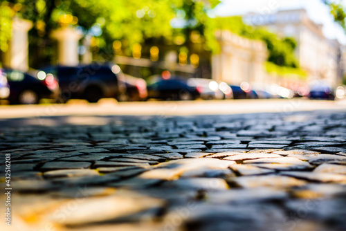 Summer in the city, the sunlit empty old street paved with stone near the park and parked cars. Close up view from the level of paving stones