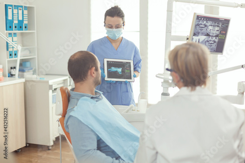 Stomatologist nurse showing tooth x-ray to sick patient explaining treatment using tablet working in stomatology hospital office. Senior doctor examining toothache while man sitting on dental chair