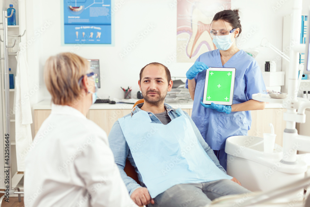 Stomatology nurse pointing her hand on mock up green screen chroma key tablet with isolated display. Sick man patient sitting on dental chair in stomatology office waiting for toothache treatment