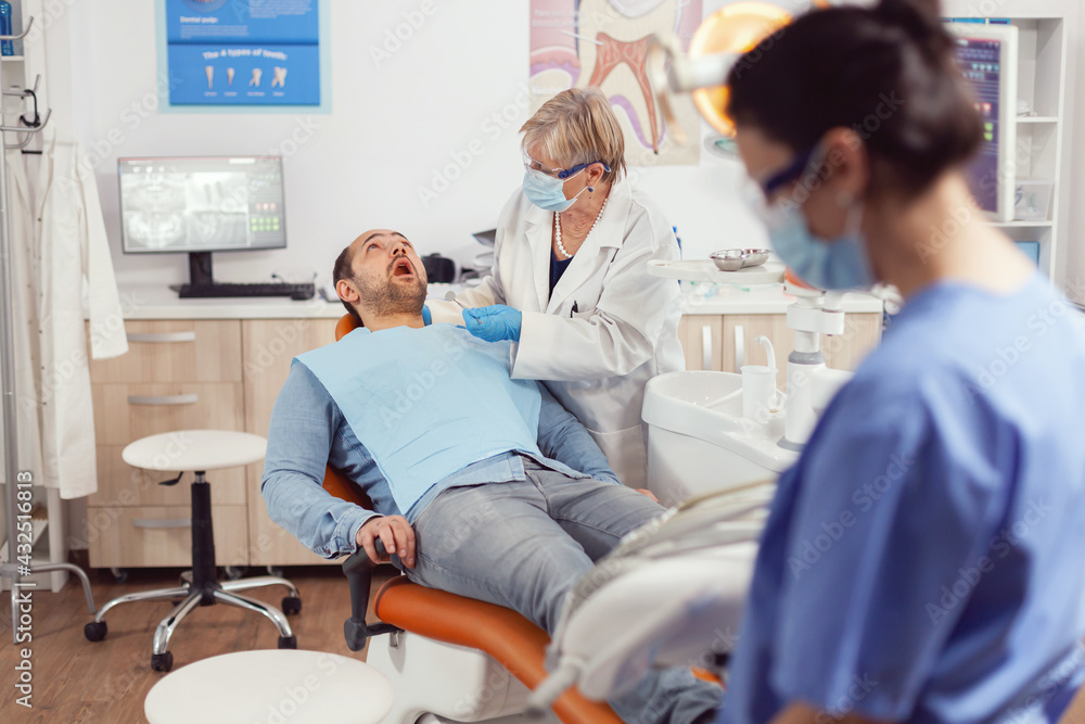 Fototapeta premium Sick patient sitting on dental chair with open mouth while senior dentist woman analyzing health teeth before medical surgery. Stomatological nurse preparing stomatology tools