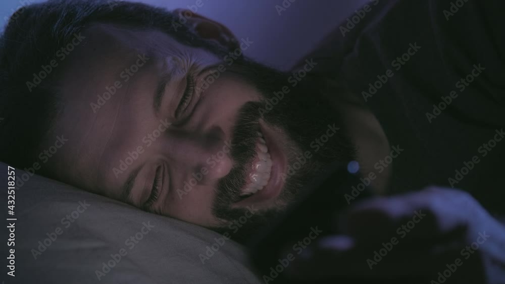Gadget night. Late chat. Internet communication. Relaxed happy smiling man enjoying reading fun joke texting message lying in bed with mobile phone in dark bedroom with blue light. Gadget night. Late chat. Internet communication. Relaxed happy smiling man enjoying reading fun joke texting message lying in bed with mobile phone in dark bedroom with blue light.