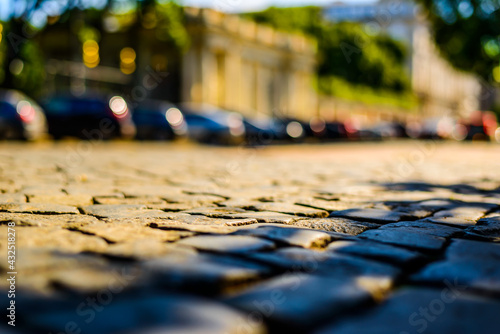 Summer in the city, the sunlit empty old street paved with stone near the park and parked cars. Close up view from the level of paving stones
