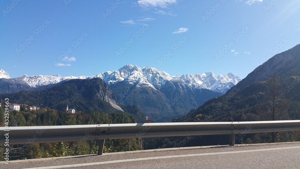 A shot on the move from the driver window of an electric car with snow-covered alps mountains in front of it in a sunny morning autumn day. POV first person view shot on a asphalted mountain road.