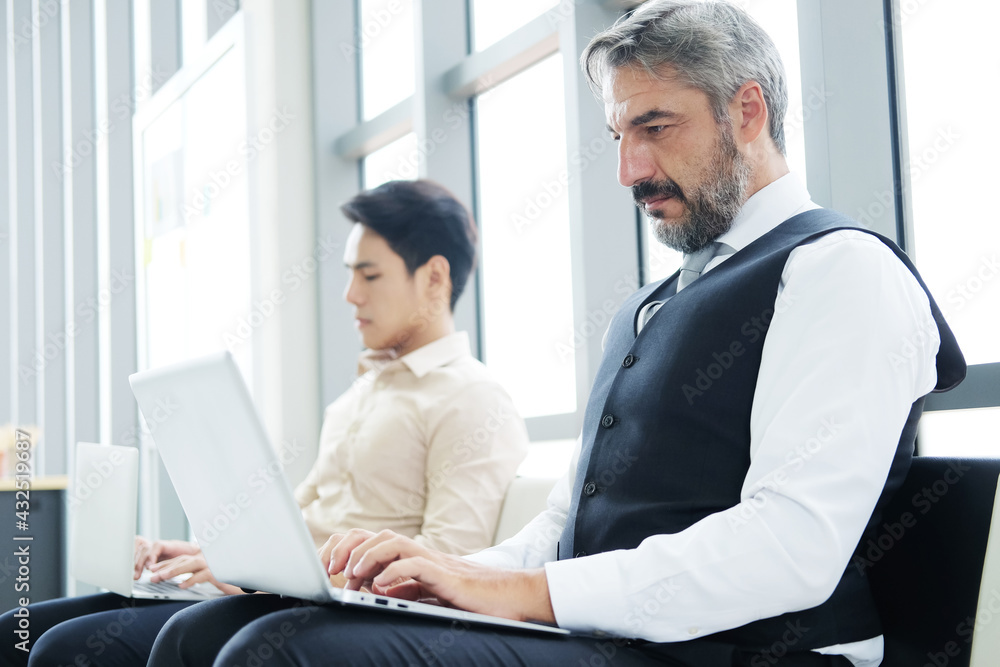 Fototapeta premium Serious Senior Caucasian businessman is working with laptop in workspace at office. He sitting distance from asian staff. Technology for business and New normal lifestyle concept.