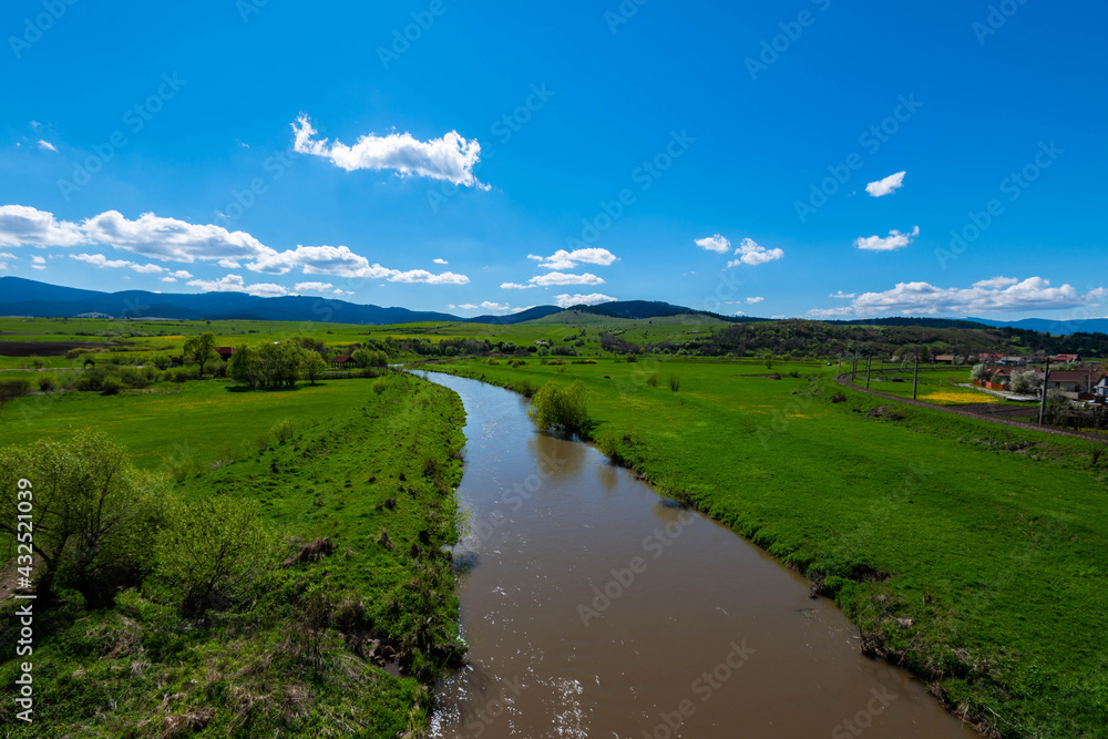 Calm flowing Olt river , vibrant green fields at spring, blue sky with white clouds.