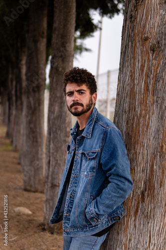 Handsome young man with curly hair and beard wearing denim posing in a public park. Half body portrait with confident expression