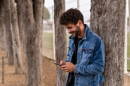 Handsome young man with curly hair and a beard looking at his smartphone and smiling in a public park with trees in the background. Technology concept