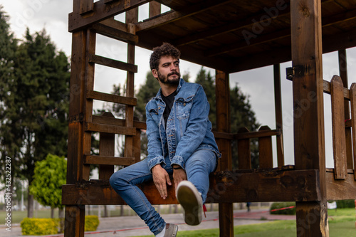 Attractive young man with curly hair and a beard wearing denim posing on a wooden playground in a public park
