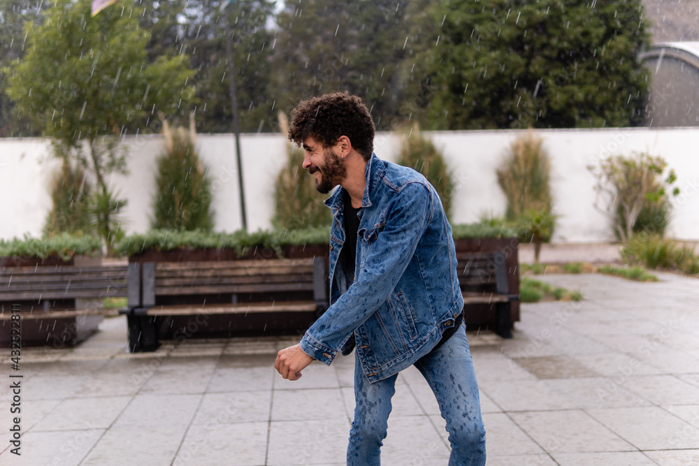 Handsome young man with curly hair and a beard wearing denim skating in the park having fun during a rainy day Handsome young man with curly hair and a beard wearing denim skating in the park having fun during a rainy day