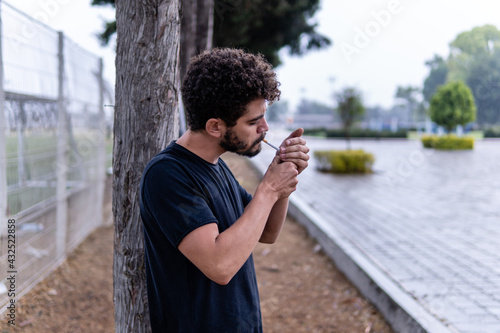 Handsome stylish young man with curly hair and a beard lighting a cigarrette up in the city park of Cholula, Mexico, with trees in the background. Smoking concept