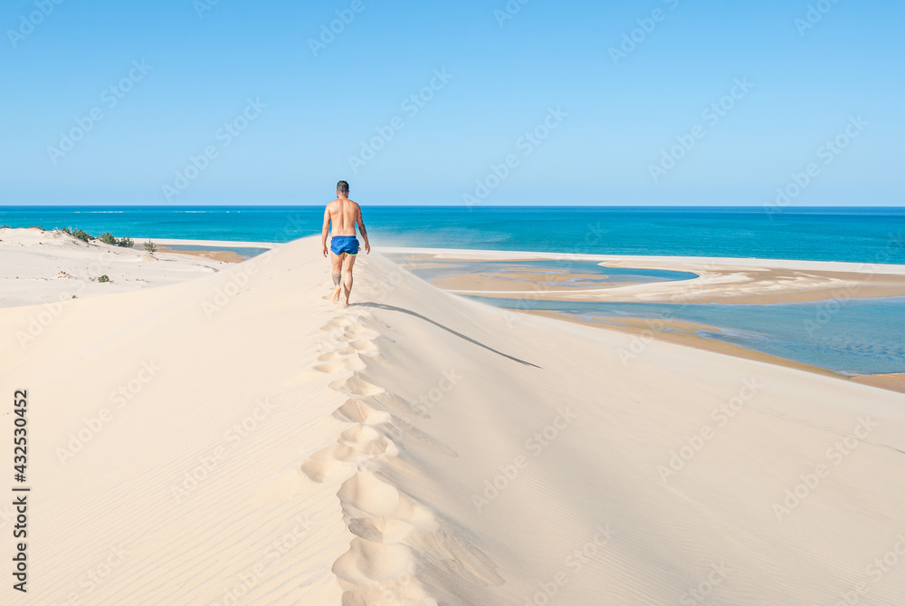 Young man walking through the sand of a dune towards the turquoise and heavenly beach of an island