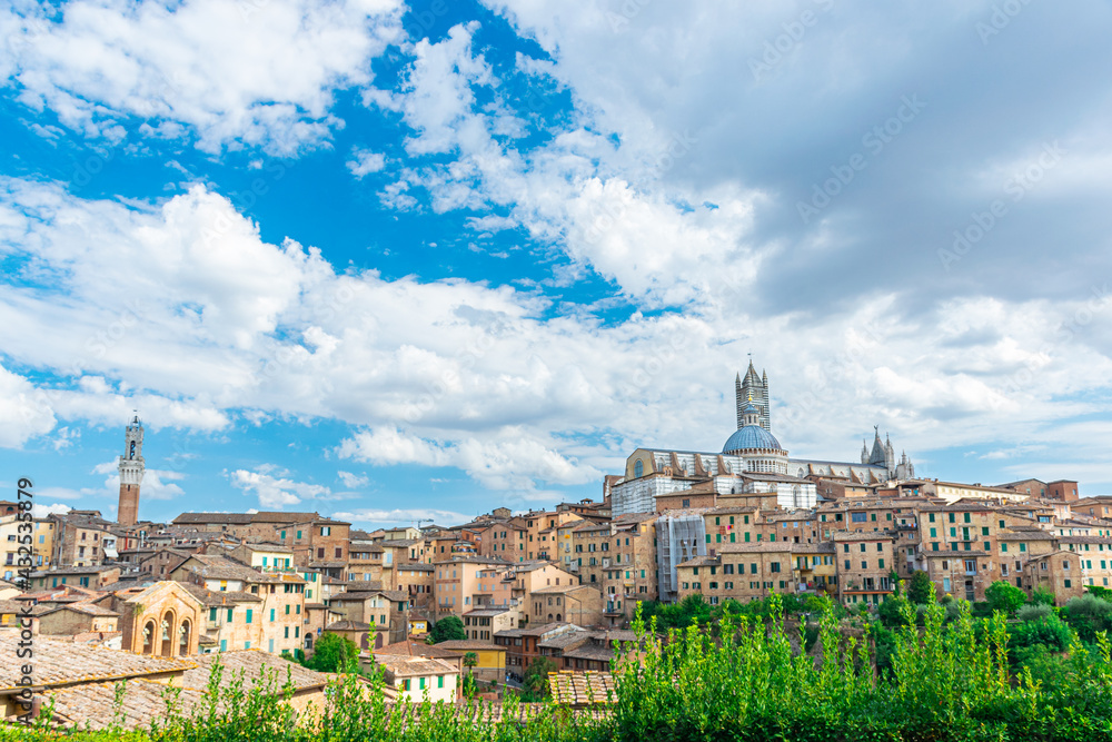 Skyline of the medieval city of Siena, with the cathedral and the city tower on a sunny day with blue sky and white clouds.