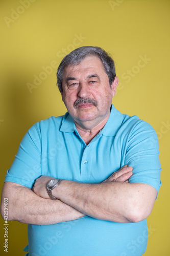 An elderly man sits on a chair and looks at the camera on a yellow background